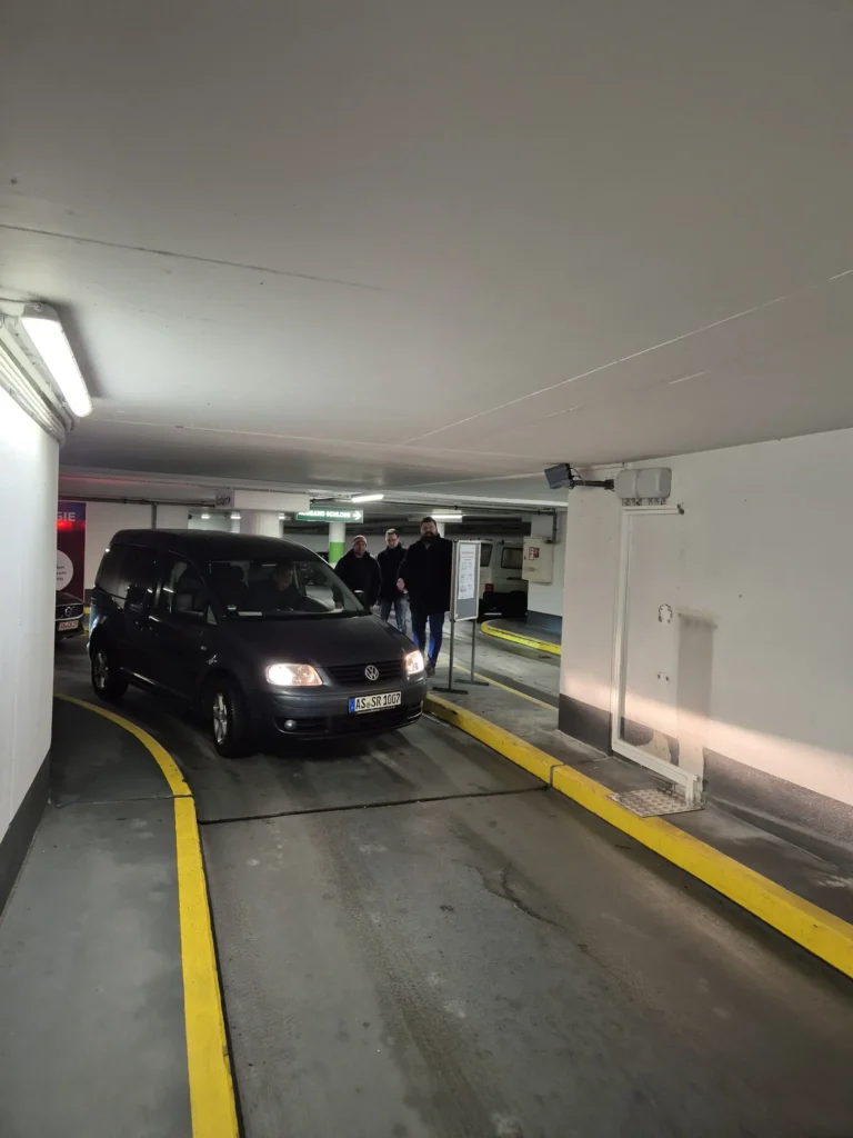 Ein Mitarbeiter fährt mit einem schwarzen VW aus der Tiefgarage Sulzbach-Rosenberg. Im Hintergrund stehen Personen nahe der Ausfahrt. Die Schranke ist geöffnet, das Fahrzeug wird per Kennzeichenerkennung erfasst.